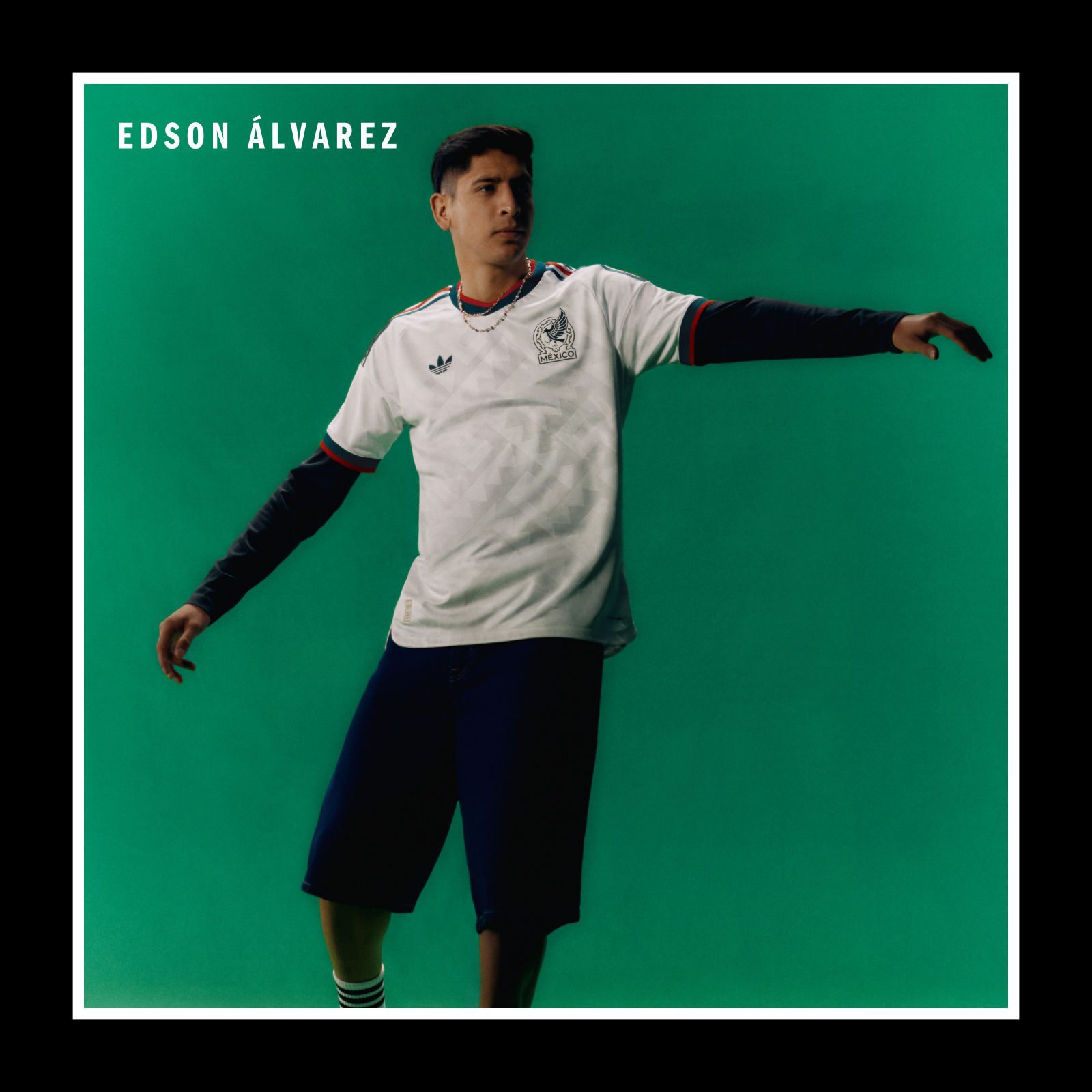 Das Bild zeigt einen jungen Mann, der ein weißes Sporttrikot mit dem Namen "Edson Alvarez" trägt und vor einem einfarbig grünen Hintergrund steht.